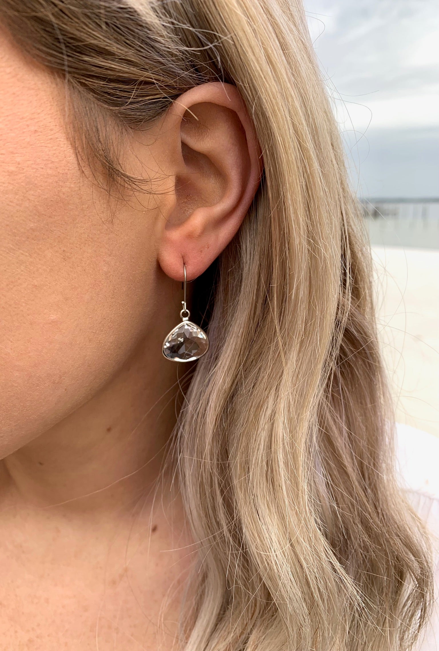Model wearing silver crystal teardrop earrings, with sand and water in the background.