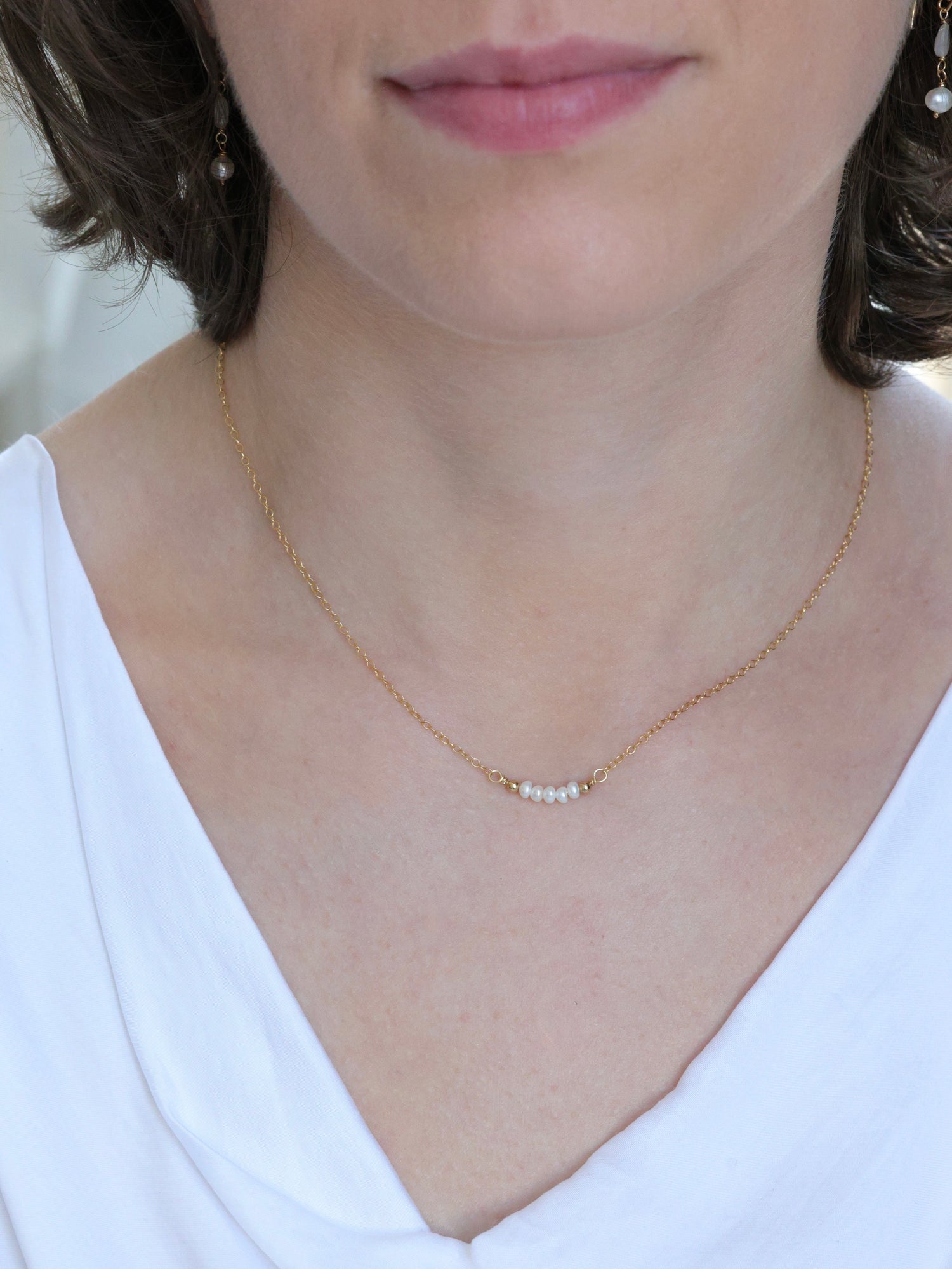 Woman wearing a minimalist gold chain necklace with a row of small white freshwater pearls at the center, styled with a white top and pearl drop earrings. Delicate and elegant look from the Nina Halls collection.