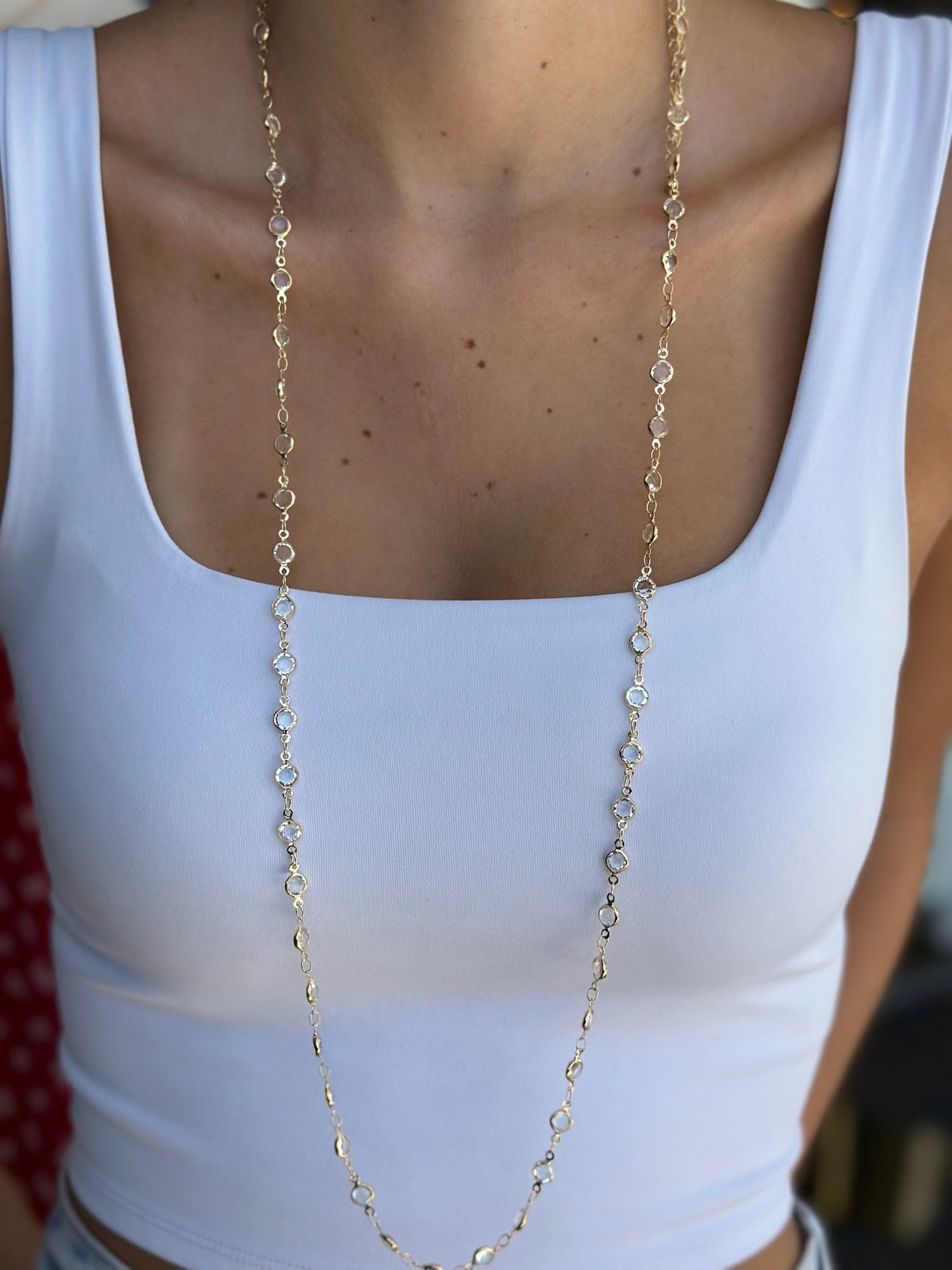 Long gold necklace worn by a person in a white tank top.