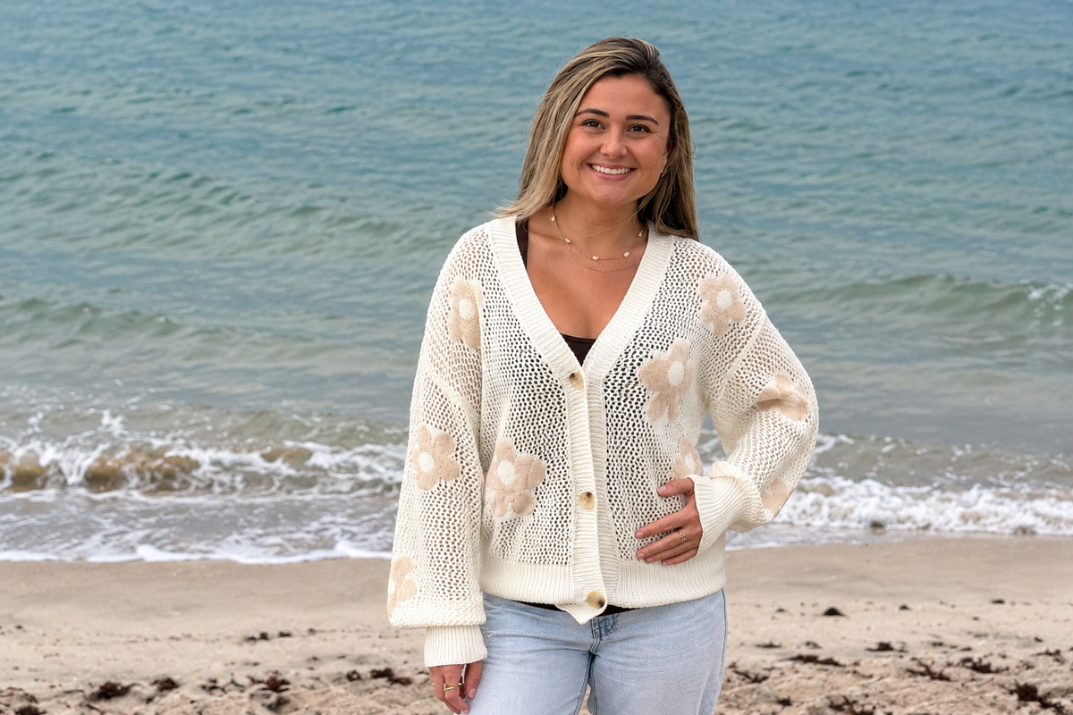 Model wearing an ivory open-knit floral cardigan with beige flowers and button-front detail by the beach at Ardor Boutique.
