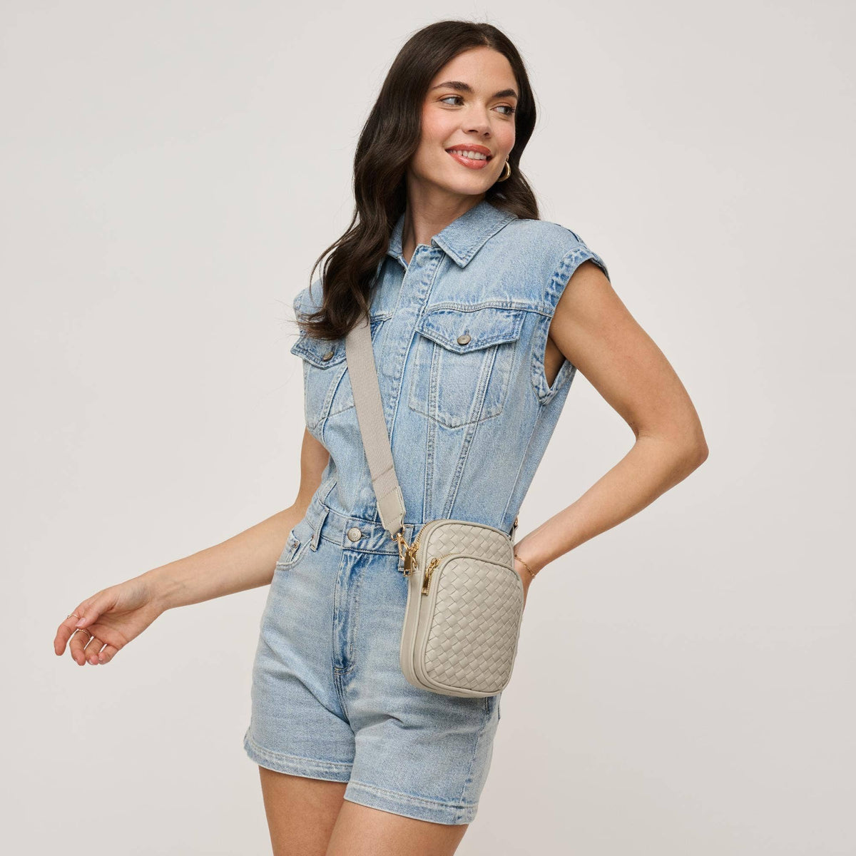A woman wearing a light blue denim romper and a beige woven crossbody bag with gold hardware, posing against a light gray studio background.
