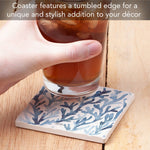 A square coaster with a blue coral pattern on a cream background, shown under a hand holding a glass of iced tea on a wooden table. Text at the top reads: 'Coaster features a tumbled edge for a unique and stylish addition to your décor'.