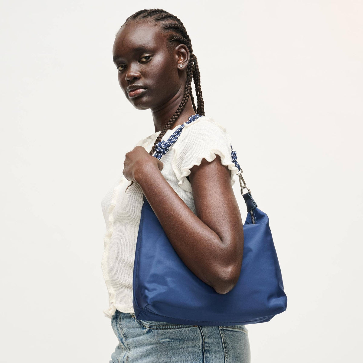 A woman with braided hair models a large blue tote bag with a blue and white patterned shoulder strap, wearing a white ruffled top and blue jeans against a plain light background.