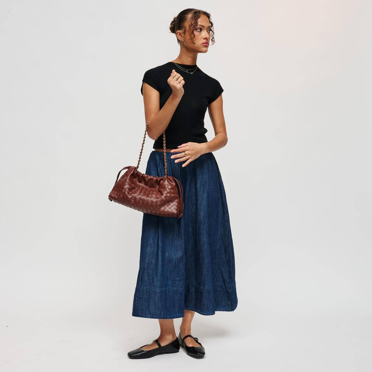 A woman stands against a plain light gray studio background wearing a black short-sleeve top, a long blue skirt, black flats, and carrying a brown woven shoulder bag.