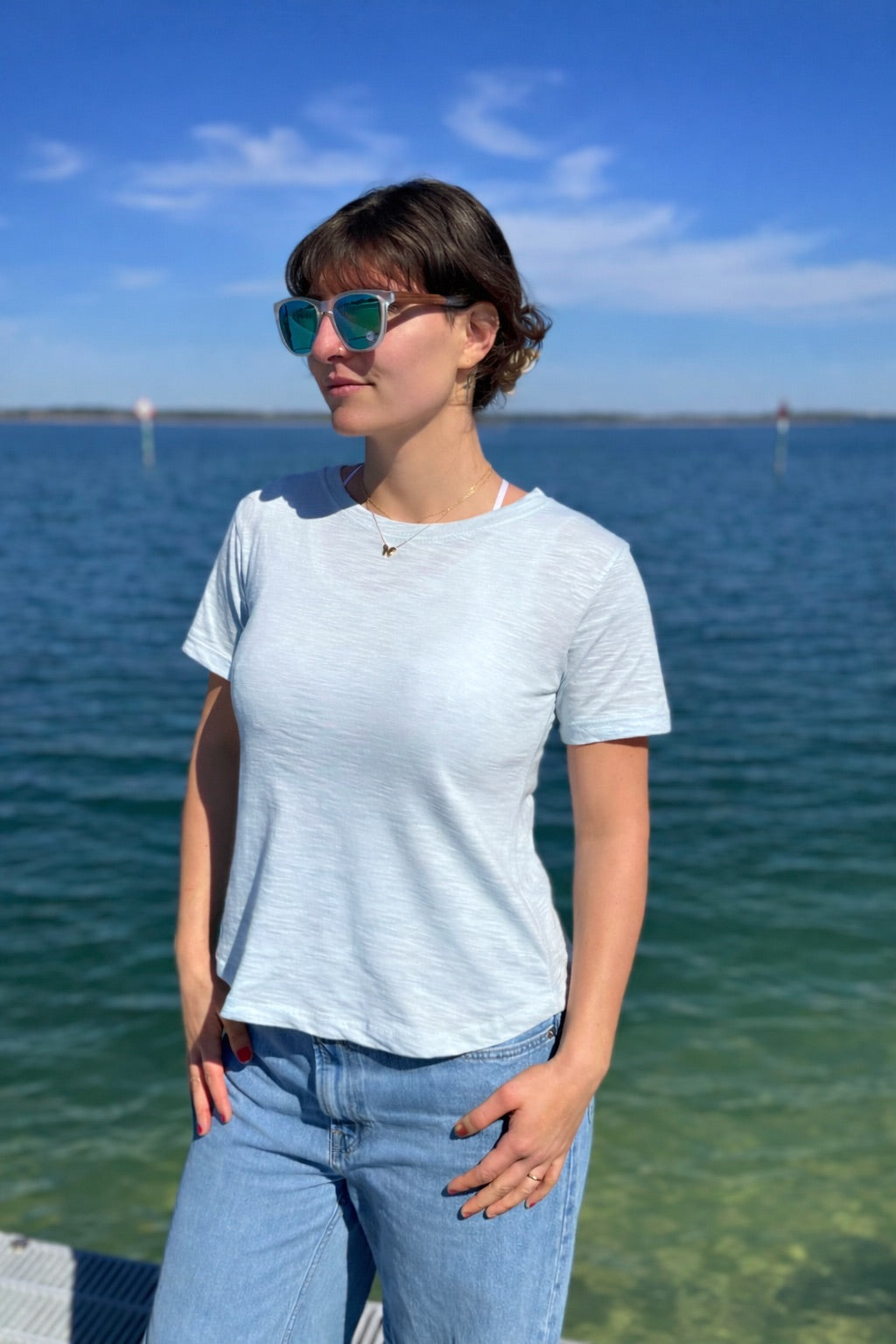 Person wearing sunglasses and a light blue shirt by a body of water