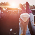 Person sitting in a car with feet up, holding a coffee cup and using a phone, sunlit interior.