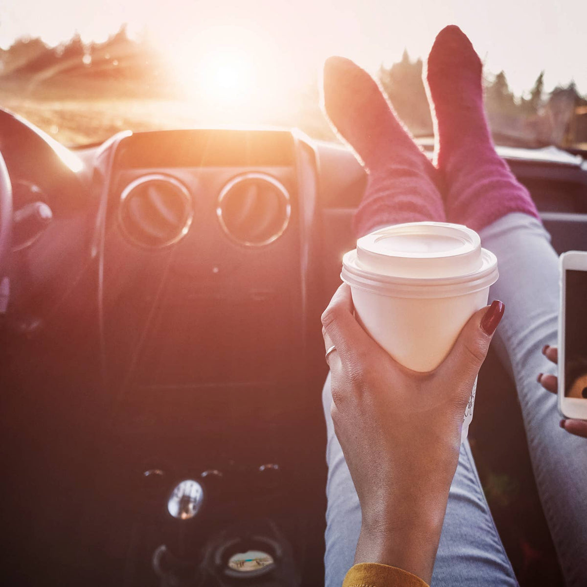 Person sitting in a car with feet up, holding a coffee cup and using a phone, sunlit interior.