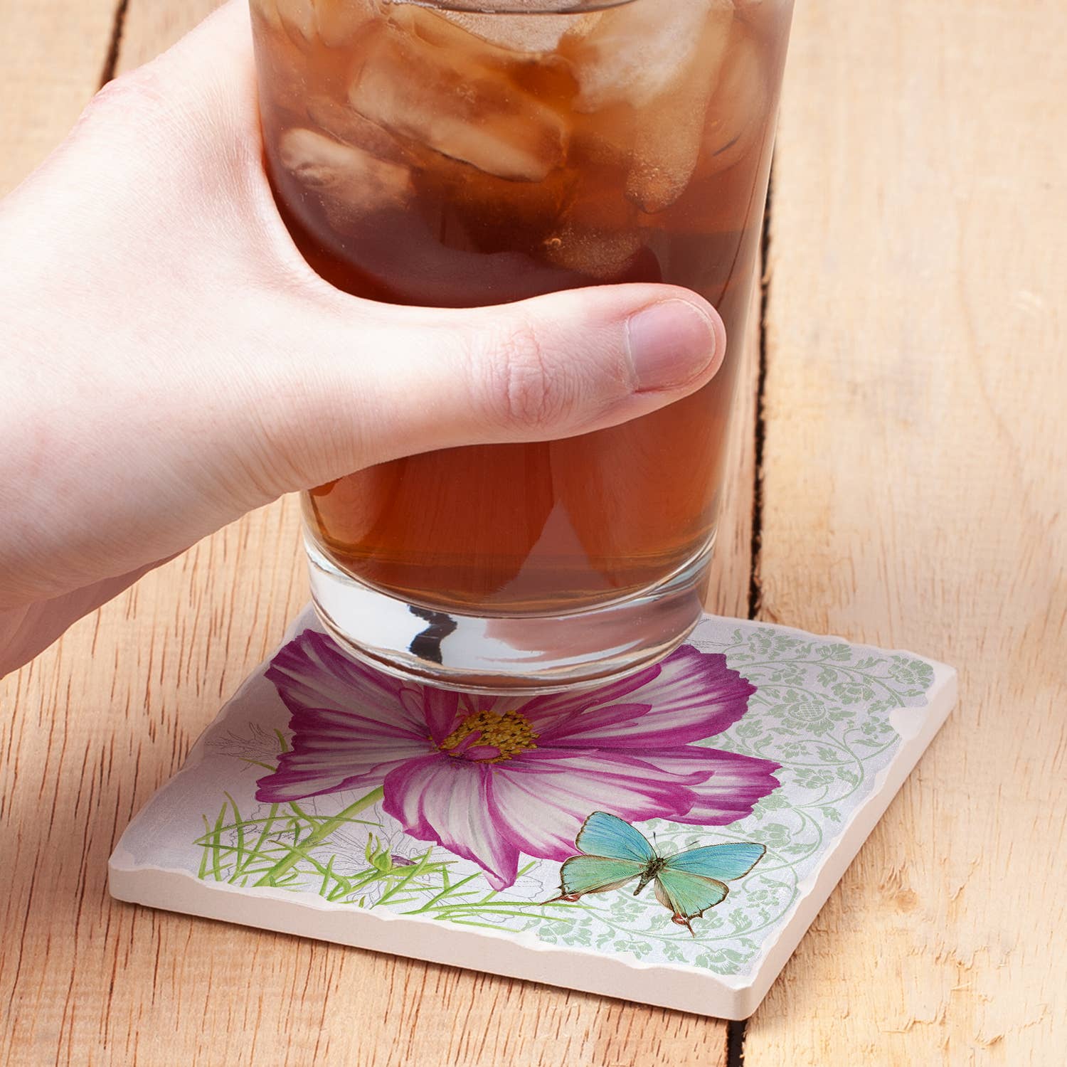 A hand holds a glass of iced coffee over a square coaster printed with a pink and purple flower, green grass, and a blue butterfly on a light wood surface.