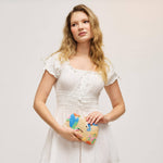 A woman with blonde hair wearing a white smocked dress and holding a small woven clutch bag with colorful bird illustrations, posed against a light gray studio background.