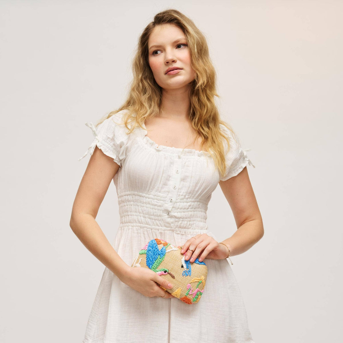 A woman with blonde hair wearing a white smocked dress and holding a small woven clutch bag with colorful bird illustrations, posed against a light gray studio background.