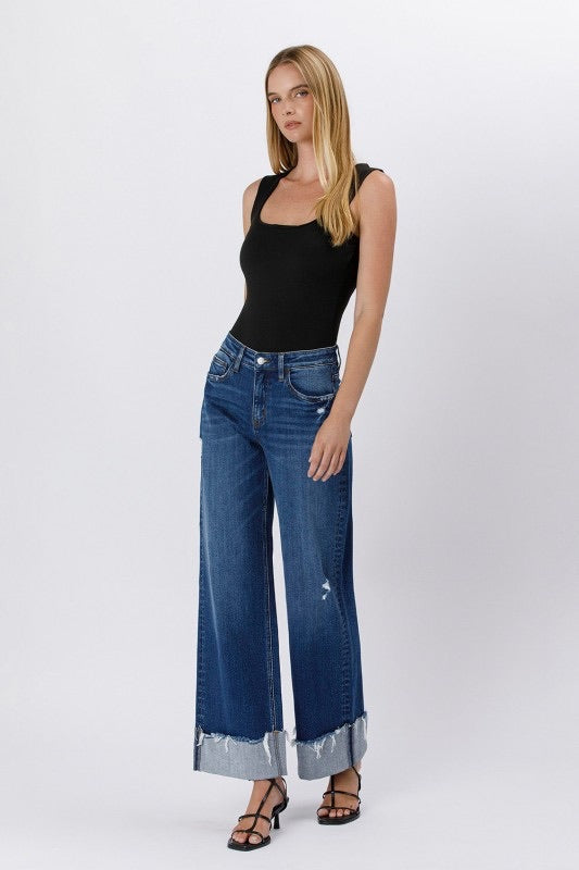 Model wearing high rise cuffed baggy wide leg jeans with slight distressing and raw single cuff hem, styled with black heels and a fitted black tank top.
