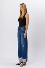 Model wearing high rise cuffed baggy wide leg jeans with slight distressing and raw single cuff hem, styled with black heels and a fitted black tank top.