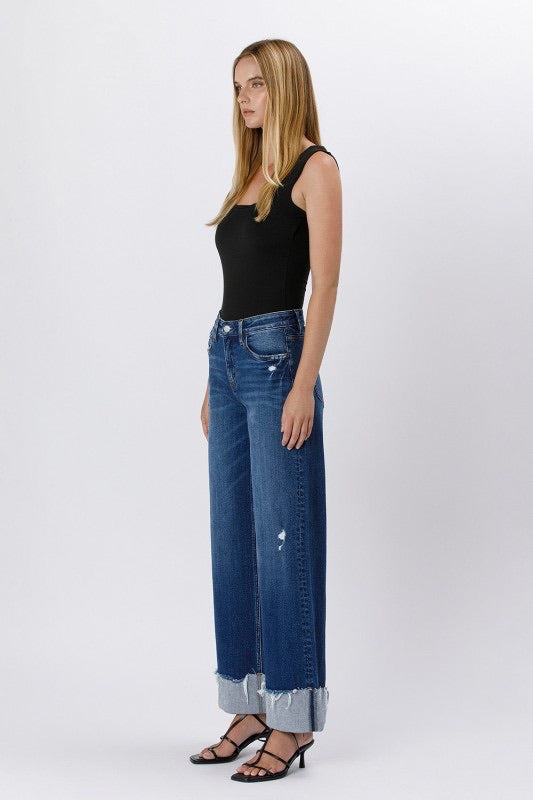 Model wearing high rise cuffed baggy wide leg jeans with slight distressing and raw single cuff hem, styled with black heels and a fitted black tank top.