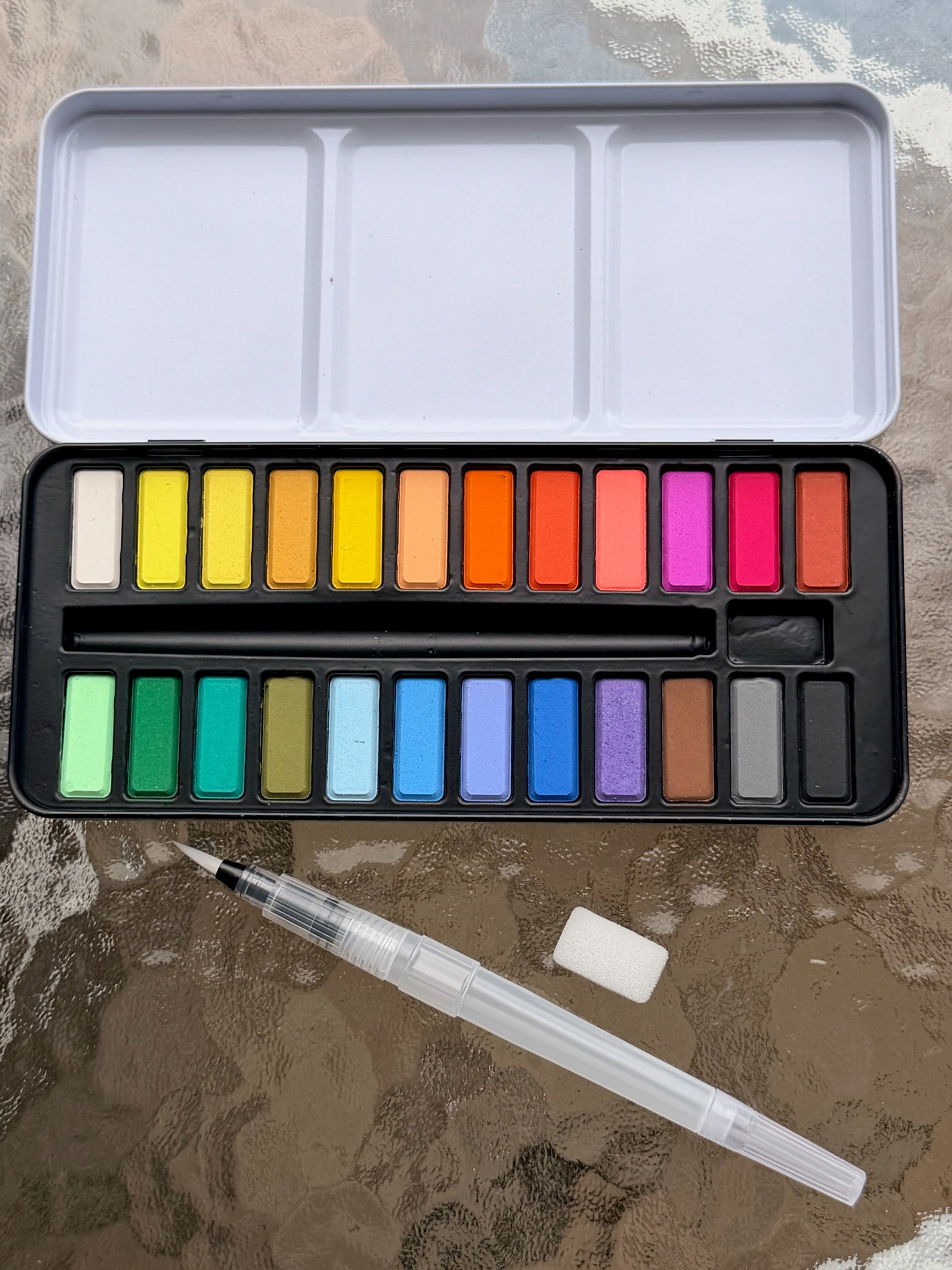 “Watercolor paint set with a range of bright and neutral colors, a refillable water brush, and sponge on a glass table.”