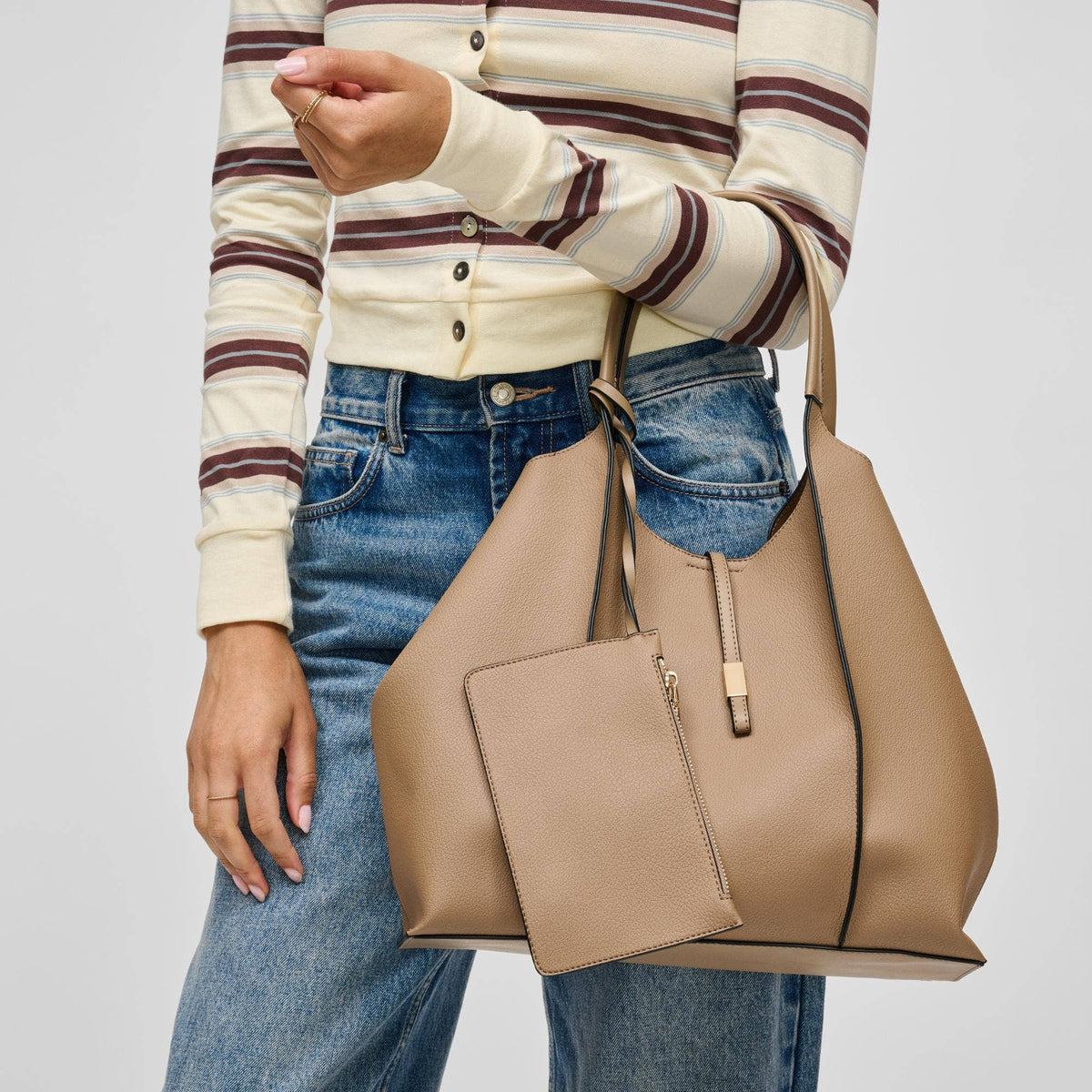 A person wearing a cream and brown striped cardigan and blue jeans carries a large taupe leather tote bag with a matching zip pouch, shown against a light gray studio background.