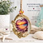 A handmade oyster shell hanging ornament with a gold glittery edge, a purple and orange sunset heart design in the center, and a small pink bead at the top. The ornament is displayed on a light wooden surface with seashells, a potted plant, and decorative text overlays.