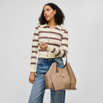 A woman modeling a beige leather tote bag with a matching zippered pouch, styled with a cream and brown striped long-sleeve top and blue jeans against a light gray studio background.