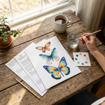 Person painting butterflies on paper with a cup of tea and art supplies on a wooden table.