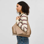 A woman in a cream and burgundy striped sweater and blue jeans carries a large taupe leather tote bag over her shoulder, shown against a light gray studio background.