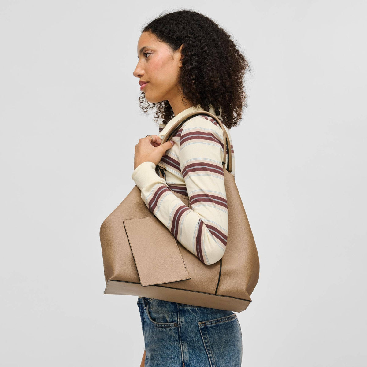 A woman in a cream and burgundy striped sweater and blue jeans carries a large taupe leather tote bag over her shoulder, shown against a light gray studio background.