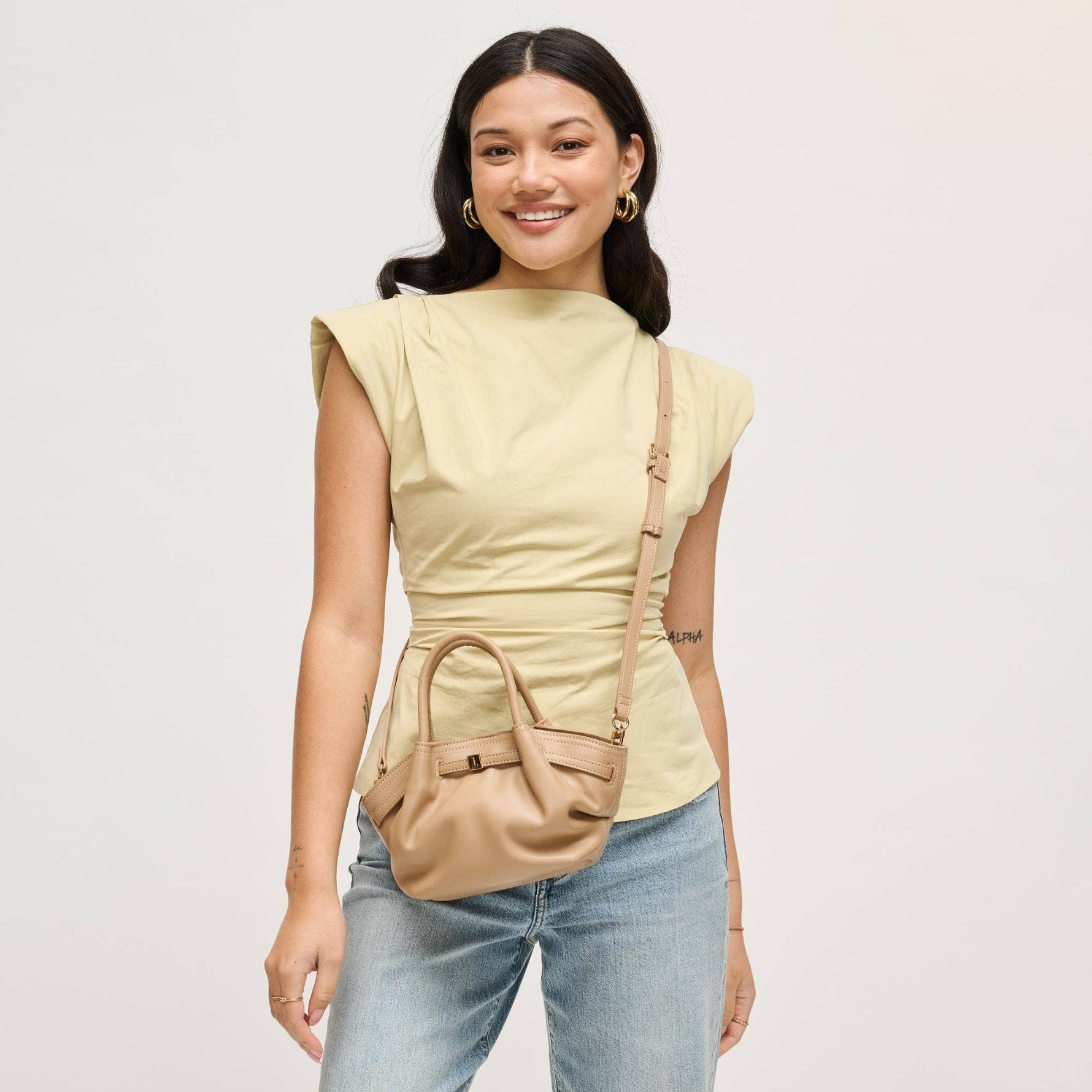 Model wearing the Selena Crossbody as a crossbody bag in taupe vegan leather with an adjustable strap and modern gathered mini satchel design.
