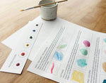 Instructional materials on watercolor techniques with a cup and brush on a wooden surface.