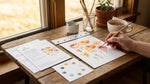Person painting flowers on paper with a brush on a wooden table near a window.