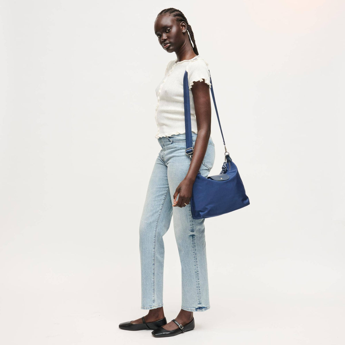 A model wearing a white ruffle-sleeve top, light-wash jeans, and black flats is shown from the side, carrying a blue shoulder bag with denim-like side panels and a blue strap against a plain white studio background.