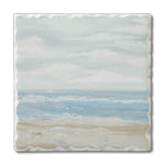 A square ceramic tile coaster featuring a painted beach and ocean scene with soft blue waves, a sandy beige shore, and a cloudy pale blue sky. The coaster has a wavy, scalloped edge and a small artist signature in the lower left corner.
