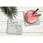 Two square coasters with a fish and coral pattern, styled on a white wood background. One coaster is empty, and the other holds a pink drink with a straw and a cucumber slice.