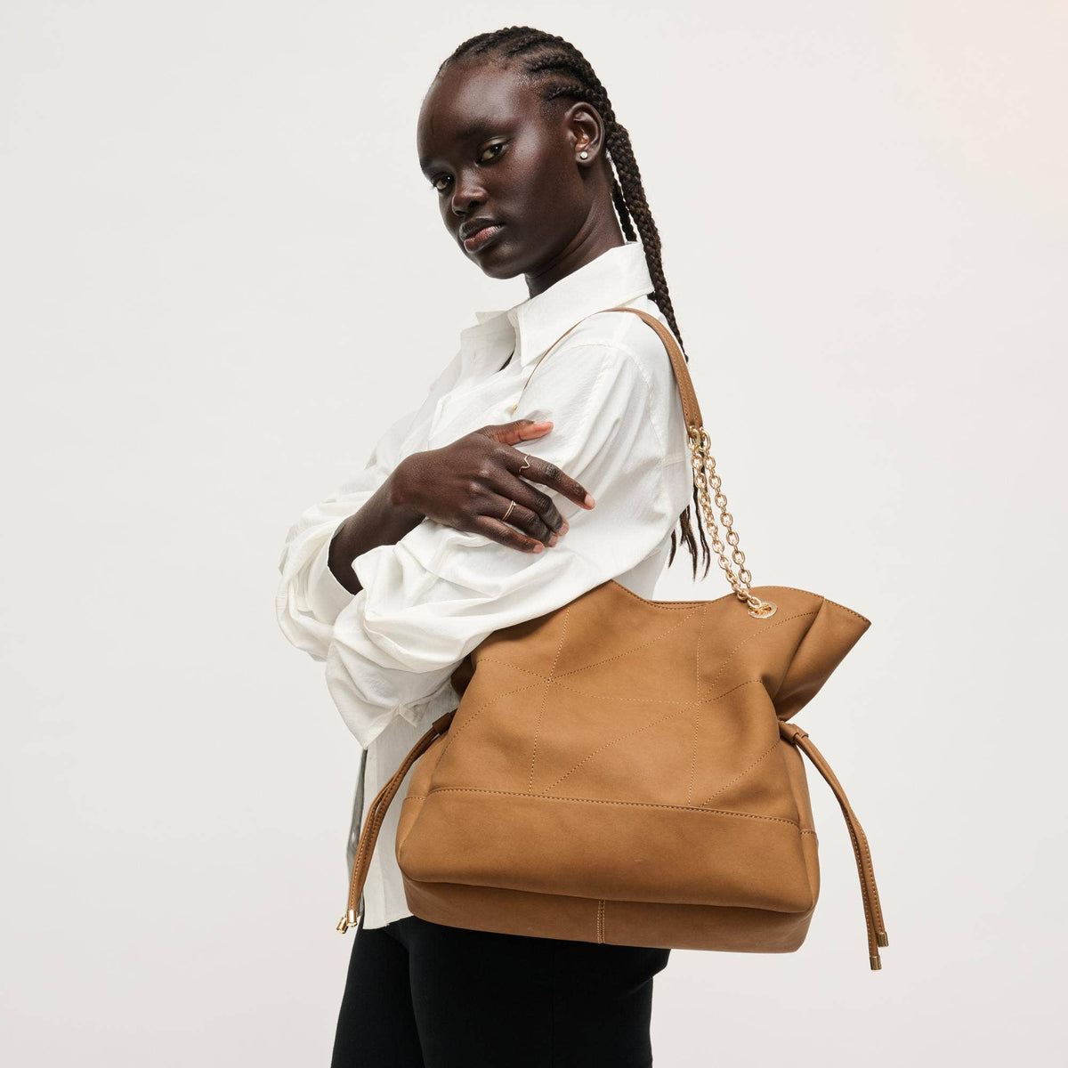 A woman wearing a white button-up shirt and black pants carries a large tan leather shoulder bag with a gold chain strap against a plain light gray background.