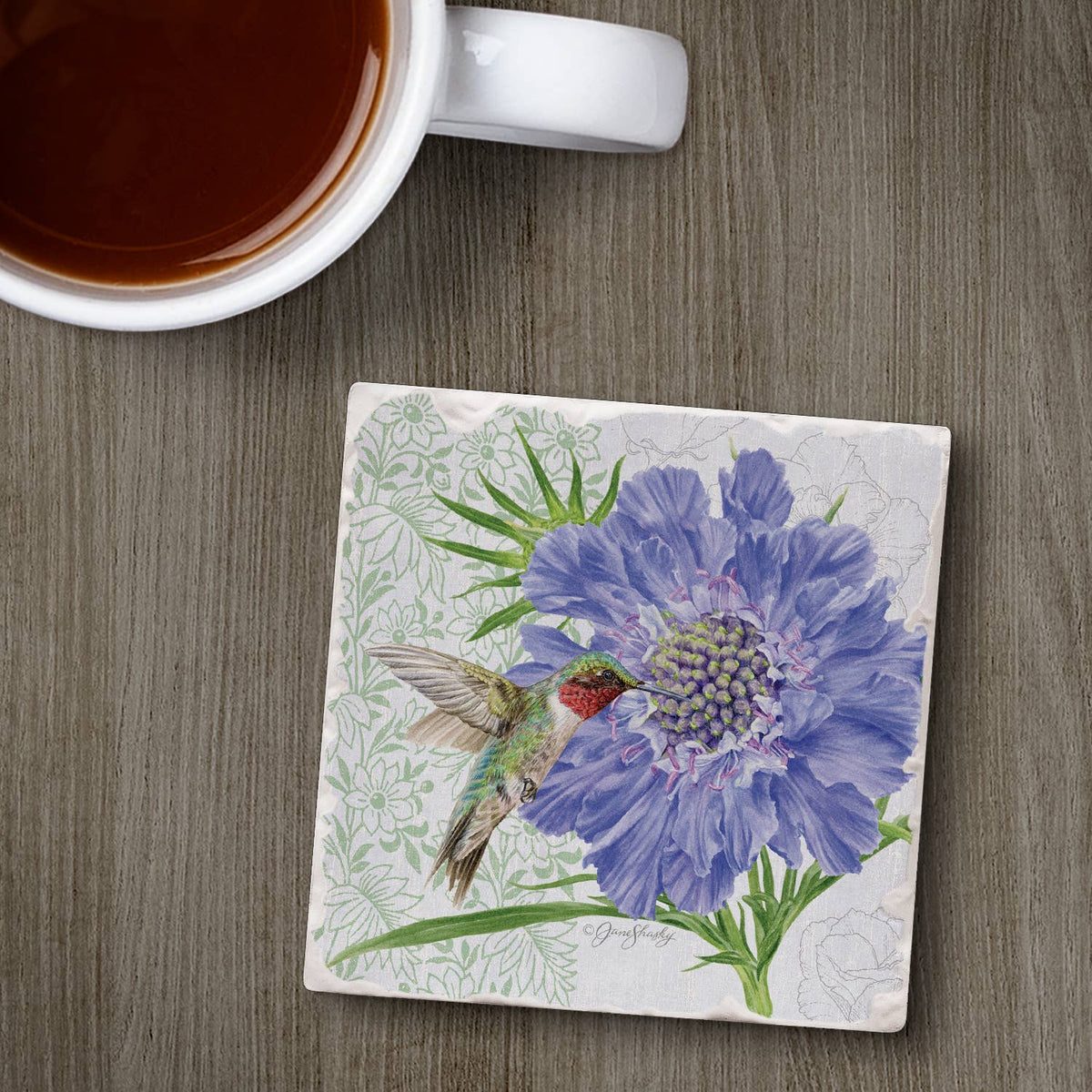 A square ceramic coaster with a detailed illustration of a blue cornflower and a hummingbird, displayed on a gray wood-grain tabletop next to a white mug filled with tea.