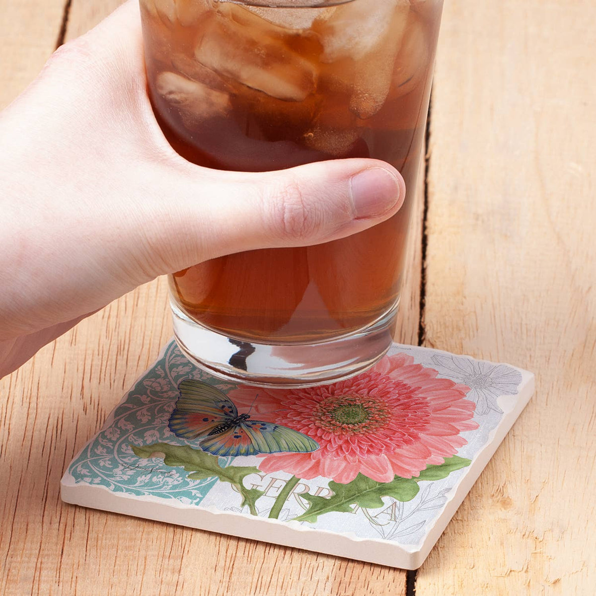Hand holding a glass of iced tea on a floral coaster with a wooden background