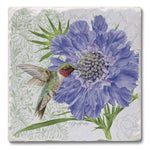 A square decorative tile or coaster featuring a painted floral design with a large blue-purple flower, green leaves, and a hummingbird on a light background with faint floral outlines.