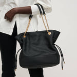 Black handbag with gold chains held by a person wearing a white shirt and black pants on a light gray background.