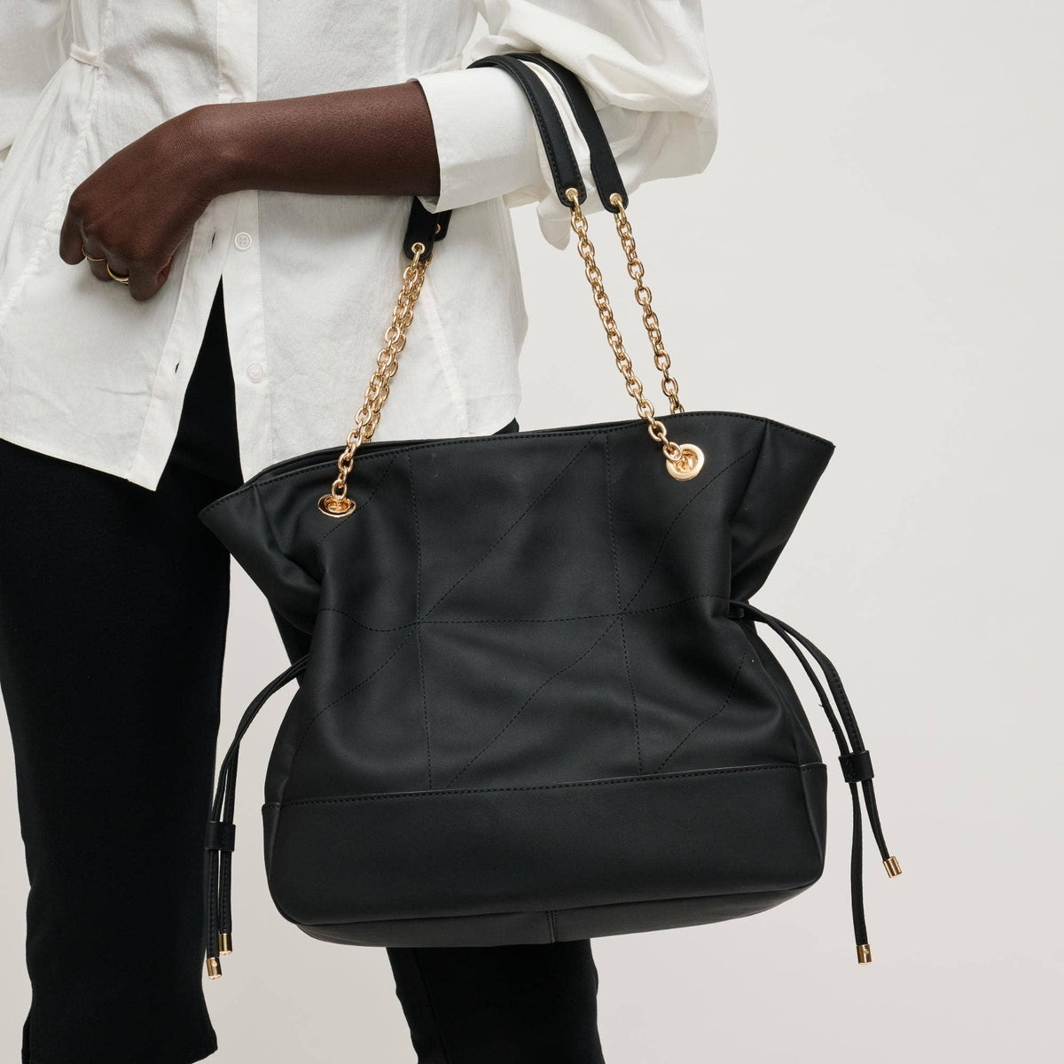 Black handbag with gold chains held by a person wearing a white shirt and black pants on a light gray background.