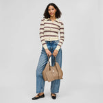 A woman stands against a light gray studio background wearing a cream ribbed knit button-front top with dark brown and burgundy horizontal stripes, blue jeans, black flats, and holding a large taupe leather tote bag.