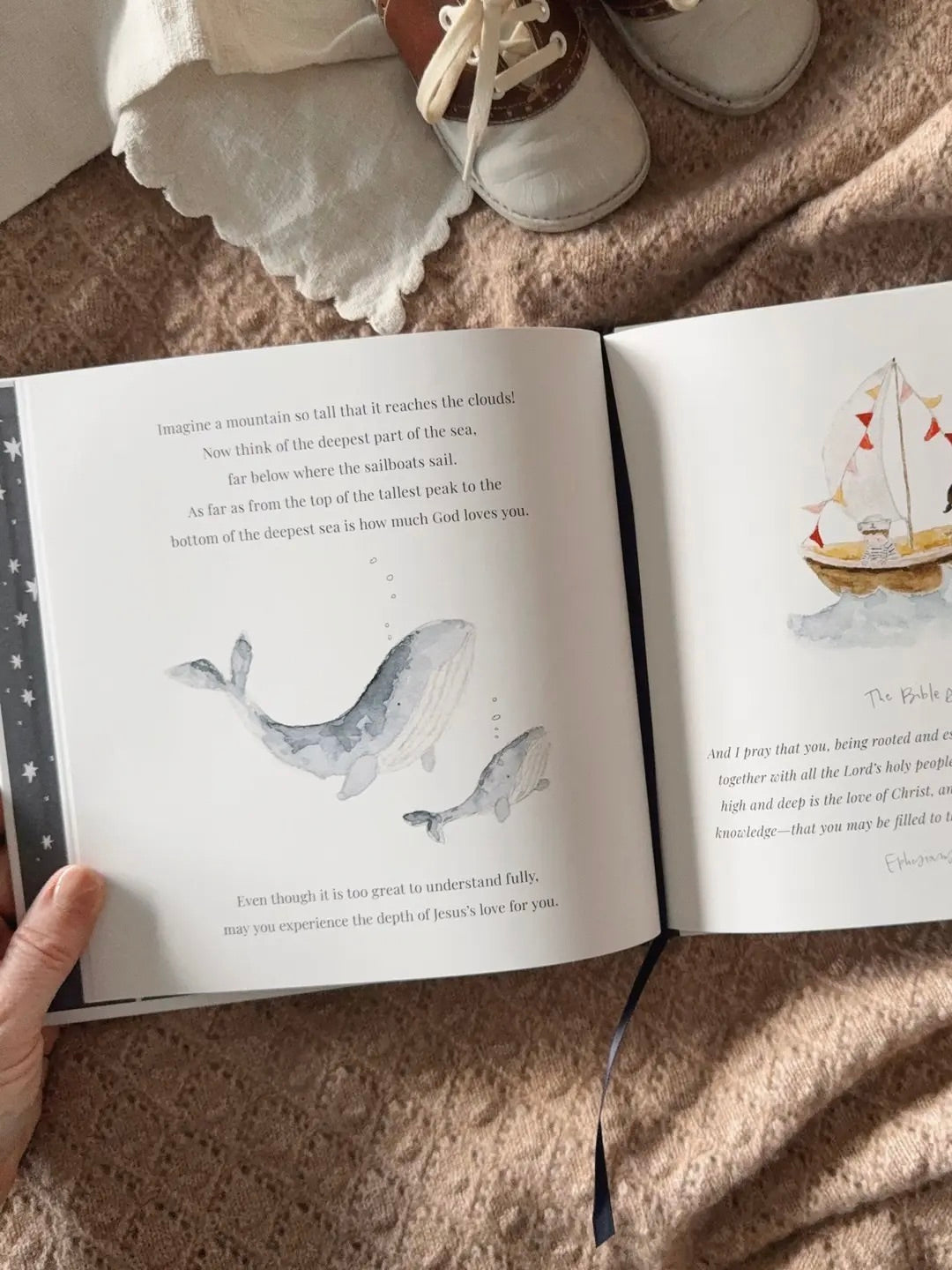 Open book with illustrations of a whale and a sailboat, held by a hand on a textured surface. Peak into book titled Bedtime Blessings. 