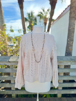 Cable knit sweater in beige on a mannequin outdoors with a long taupe necklace