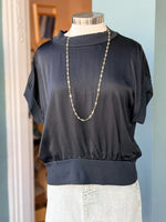 Black blouse on a mannequin with a gold chain necklace against a light blue wall.