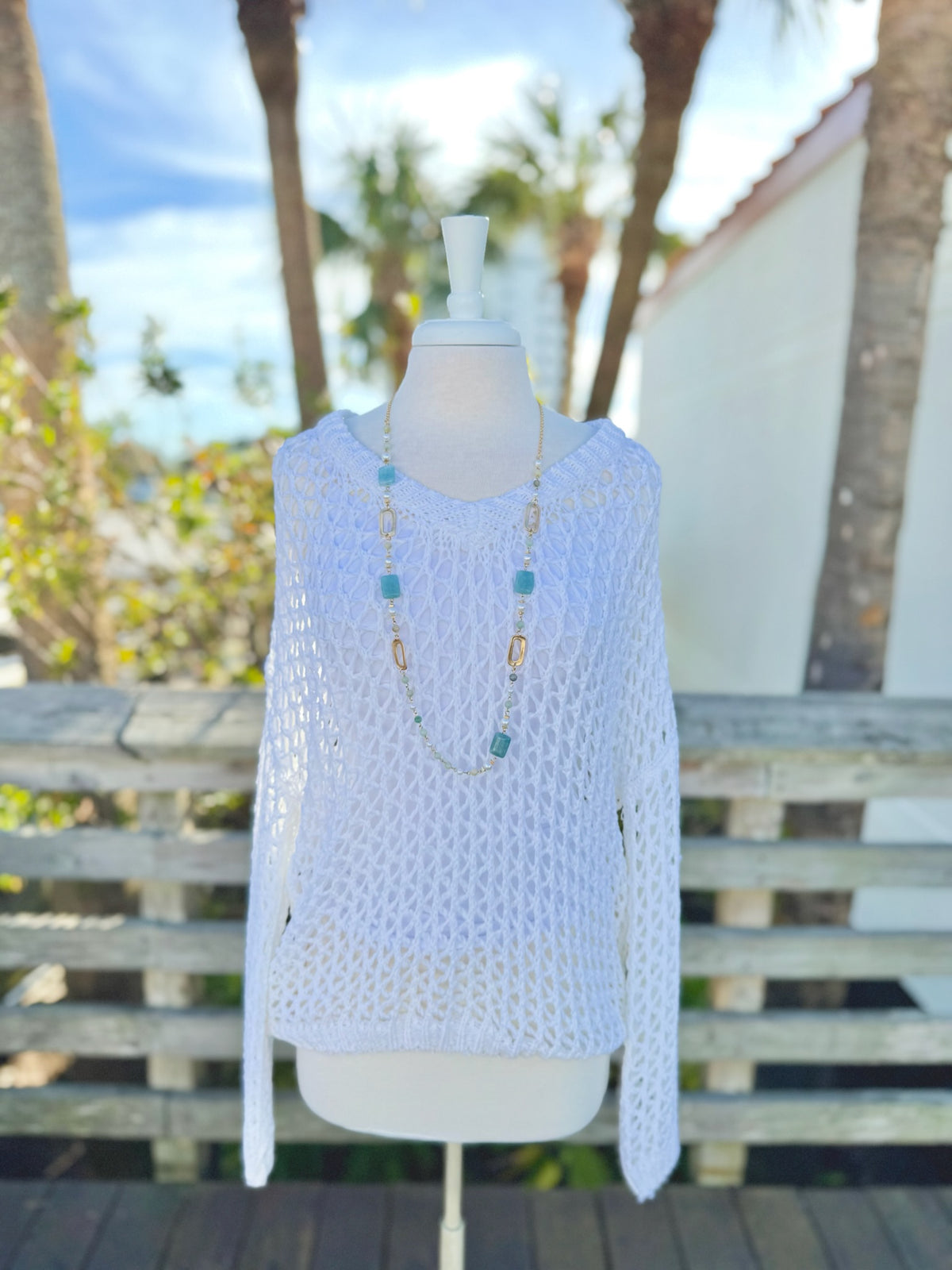 White open knit sweater on a mannequin outdoors with a long blue and gold necklace