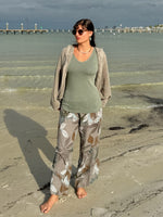 Model standing barefoot on the beach wearing the Catherine Linen Pants in mocha floral with an olive tank and taupe mesh jacket, waves in the background.