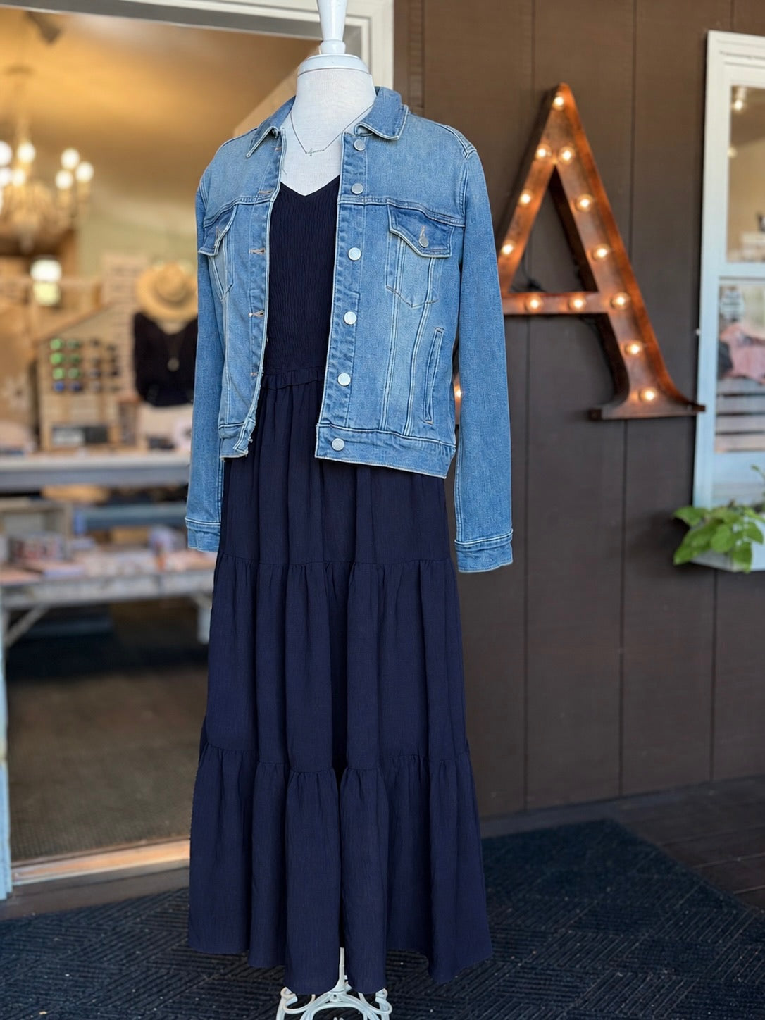 Navy maxi dress with a smocked bodice, V-neckline, ruffle short sleeves and a flowing tiered skirt paired with a jean jacket at Ardor Boutique.