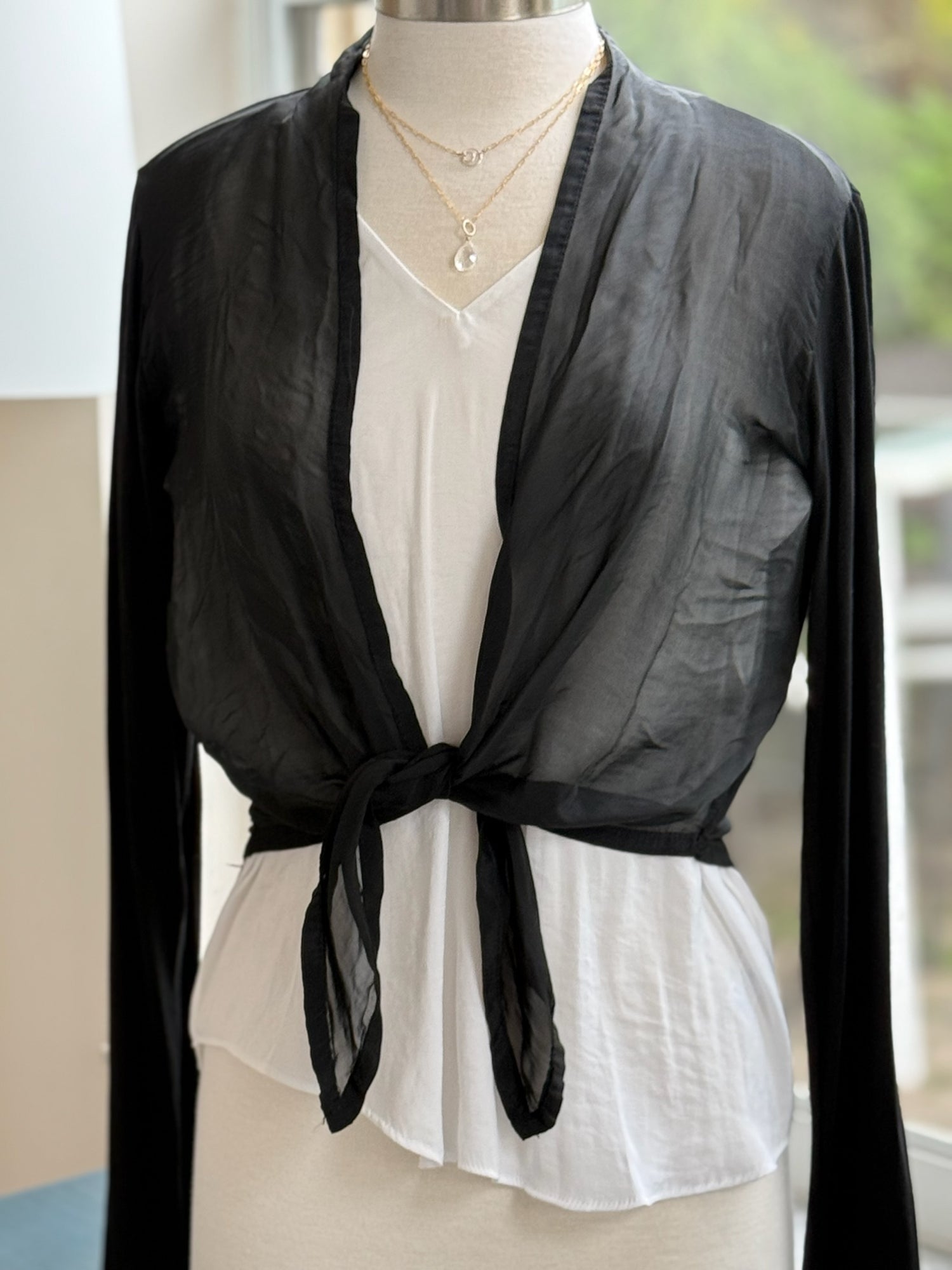 A black silk cardigan with a tie at the waist, displayed on a mannequin that is wearing a white top and a necklace.