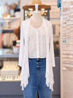 A white lace-trimmed long-sleeve cardigan layered over a white lace blouse and paired with a blue denim skirt, displayed on a mannequin torso inside a retail store.