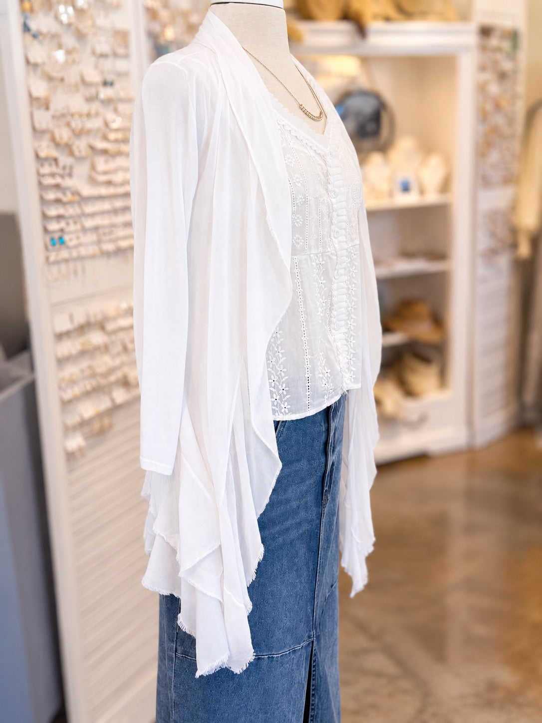 A white lace-trimmed long-sleeve cardigan layered over a white lace blouse and paired with a blue denim skirt, displayed on a mannequin torso inside a retail store.