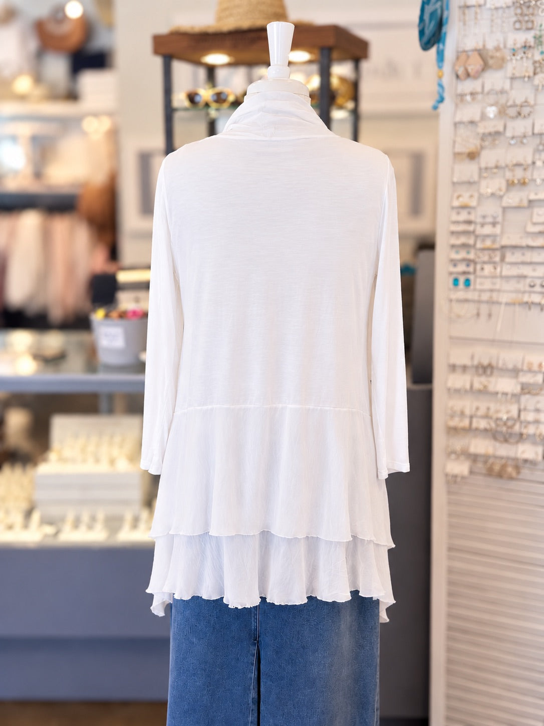 Back view of a  white lace-trimmed long-sleeve cardigan layered paired with a blue denim skirt, displayed on a mannequin torso inside a retail store.