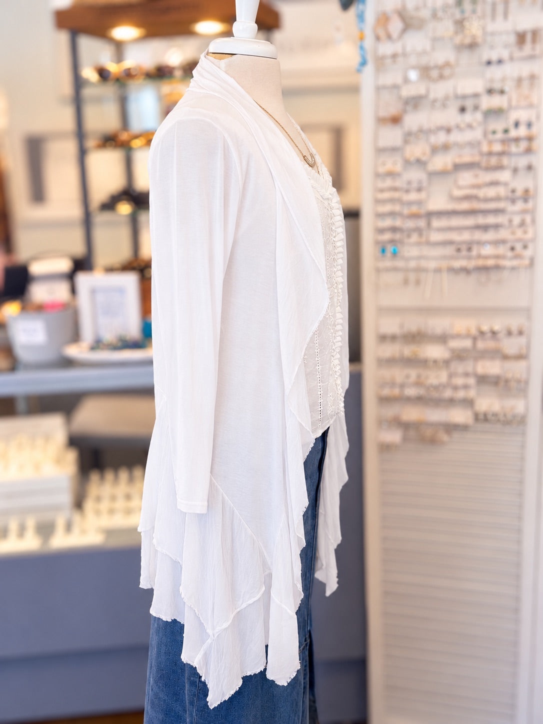 Side view of a white lace-trimmed long-sleeve cardigan layered over a white lace blouse and paired with a blue denim skirt, displayed on a mannequin torso inside a retail store.