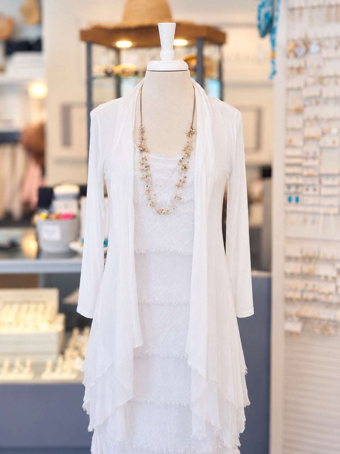 A white tiered lace dress displayed on a mannequin in a jewelry store, styled with a long beaded necklace and a sheer white draped cardigan.
