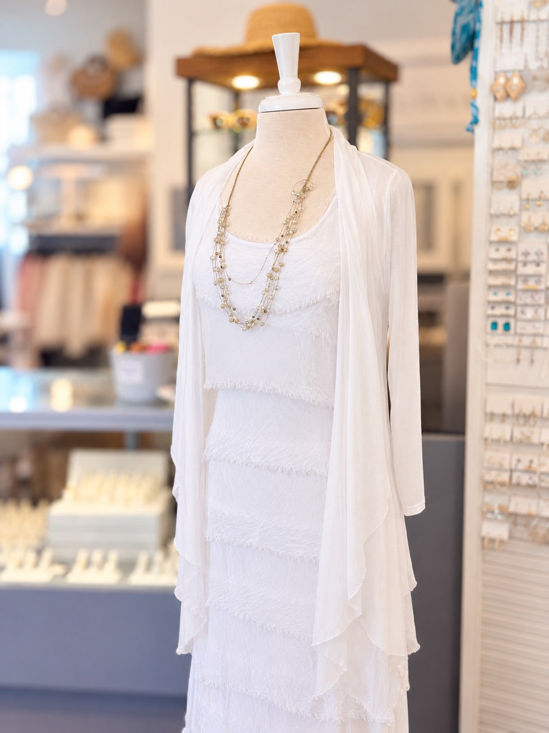 A white tiered lace dress displayed on a mannequin in a jewelry store, styled with a long beaded necklace and a sheer white draped cardigan.